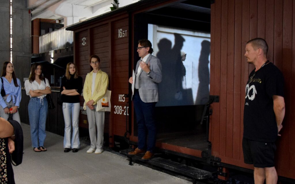 One person talking to a group of people in one of the rooms of the museum. On the background a wagon of a train exposed in the exhibition.